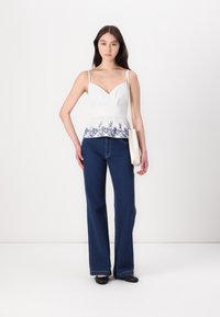 Witte peplum top met marineblauwe bloemenborduursels, gecombineerd met donkerblauwe flared jeans. Het model draagt ook een lichtgekleurde tote bag.
