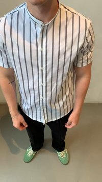 Camisa de botão de mangas curtas em branco com riscas verticais pretas, combinada com calças pretas e sapatilhas verdes com detalhes brancos.
