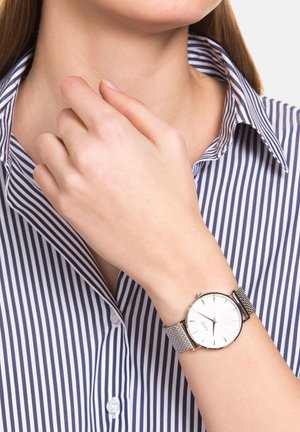 Silberner Mesh-Uhrenarmband mit weißem rundem Ziffernblatt und silbernen Stundenmarkierungen, am Handgelenk getragen, kombiniert mit einem navy und weiß gestreiften Hemd.