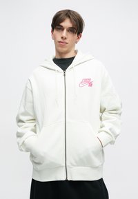 Sweatshirt branco com fecho éclair, feito de tecido macio, apresentando um logótipo da Nike em bordado rosa no peito e bolsos frontais.