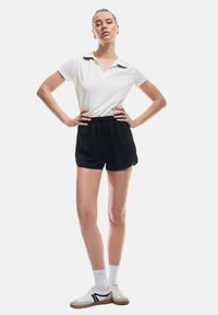 Camiseta polo blanca con cuello negro, combinada con pantalones cortos negros. El modelo lleva zapatillas blancas con acentos negros y calcetines blancos.