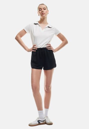 Camiseta polo blanca con cuello negro, combinada con pantalones cortos negros. El modelo lleva zapatillas blancas con acentos negros y calcetines blancos.