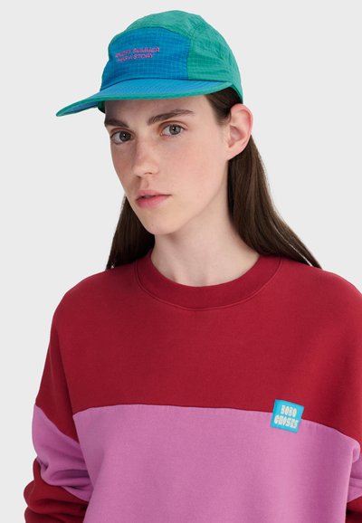 Jeune personne aux longs cheveux bruns portant une casquette bleu sarcelle et bleu avec l'inscription "CHAQUE ÉTÉ A UNE HISTOIRE" ainsi qu'un sweat-shirt rouge et rose avec un écusson bleu.