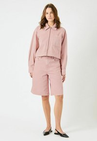 Kvinne iført en lys rosa jakke med glidelås og matchende shorts til kneet, kombinert med svarte spisse slingback-sko, stående på hvit bakgrunn.