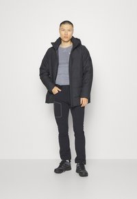 Giacca a vento nera con cappuccio, camicia grigia aderente sotto, pantaloni neri slim-fit e scarpe da trekking nere. Tessuto liscio con motivo trapuntato.