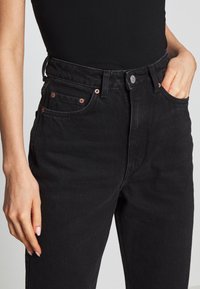 Czarne dżinsy w stylu denim z wysokim stanem, z mosiężnymi detalami, przednimi kieszeniami i lekko teksturowanym materiałem.