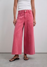 Pantaloni rosa a gamba larga in denim, con vita alta, tasche laterali e cuciture a contrasto. Abbinati a sandali neri.