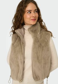 Beige faux fur vest met een opstaande kraag en een ritssluiting aan de voorkant. Voorzien van zijzakken met rits en een zachte binnenvoering. Gestructureerde, gezellige uitstraling.
