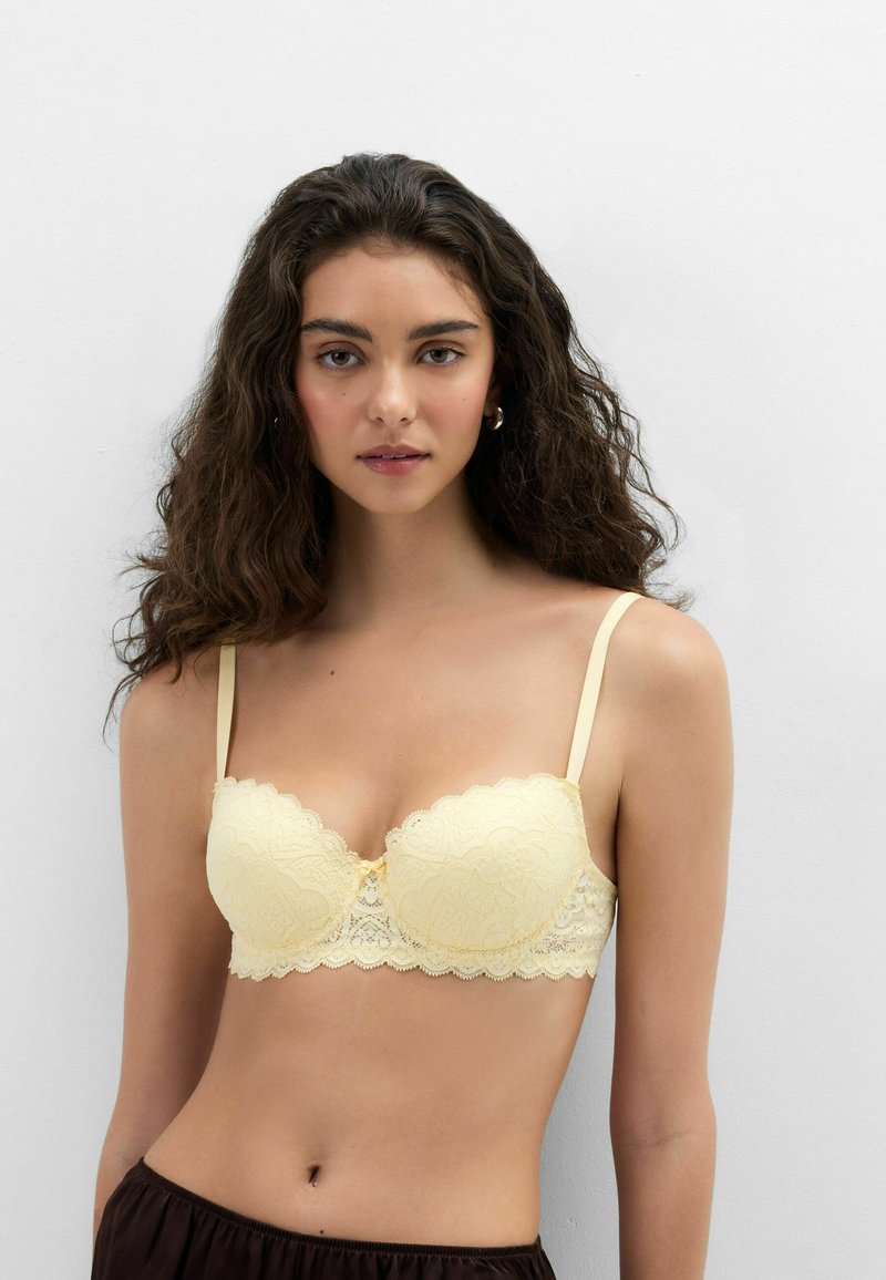 Woman with long curly hair wearing a light yellow lace bra and dark bottoms against a plain white background.