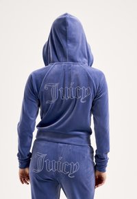 Mélykék bársony melegítő felső kapucnival. A hátán csillogó strasszkövekkel díszített "Juicy" felirat, sima textúra és testhezálló kialakítás.