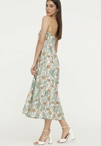 Blumenkleid ohne Träger mit einem grün-weißen Muster und orangefarbenen Blumen. Der Saum ist ausgestellt und wird mit weißen Blockabsatzsandalen kombiniert.