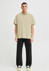 Beige polo skjorte med korte ermer, krage, løs passform, kombinert med svarte vide bukser og sneakers med hvite og svarte detaljer.