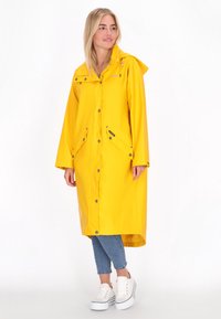 Hosszú sárga esőkabát kapucnival, patent gombokkal és elülső zsebekkel. Simább, vízálló textúrával rendelkezik, és ívelt alsó szegéllyel készült.