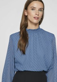 Niebieska bluzka z długim rękawem, o teksturowanym wzorze w zygzak, z falbanistym wysokim kołnierzem i półprzezroczystym materiałem, zestawiona z czarnym dołem.