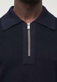 Tmavomodré polo tričko so zipsom, s rebrovanou textúrou, plochým golierom a strieborným zipsom. Má krátky dizajn krku a nenápadné logo na ťaháku zipsu.