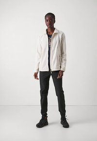 Krema puhasta flis jakna z visokim ovratnikom in zadrgo, nošena čez temno majico, kombinirana s črnimi hlačami in športnimi čevlji.