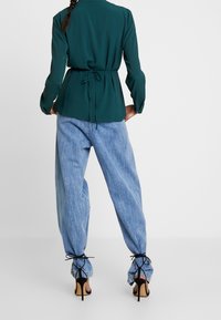 Grön texturerad blus med cinchad midja, i kombination med ljusblå baggy-jeans med knytdetaljer vid anklarna. Svarta högklackade skor är synliga.