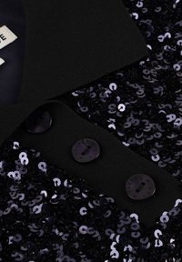 Zwarte textuur jas kraag met drie ronde knopen en dicht op elkaar geplaatste glanzende zwarte pailletten op de rand van de stof.