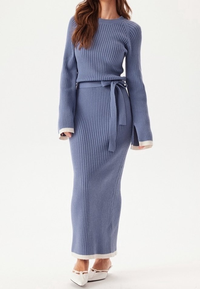 Femme portant une longue robe en maille bleue côtelée avec un lien à la taille et une bordure blanche, assortie à des chaussures blanches à bout pointu.