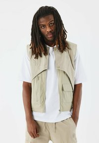 Gilet senza maniche di un beige chiaro, con zip e chiusure a pressione, tasche frontali, design a strati, indossato sopra una maglietta bianca. Tessuto morbido e leggero.