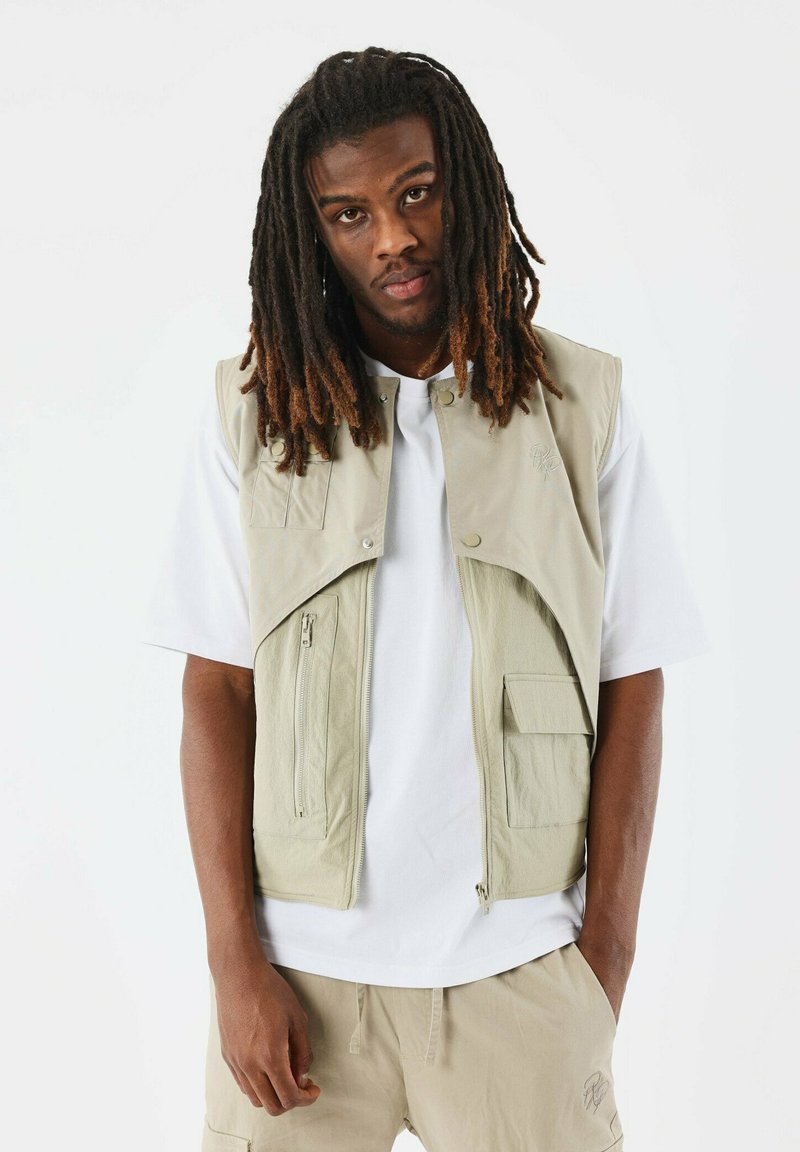 Gilet senza maniche di un beige chiaro, con zip e chiusure a pressione, tasche frontali, design a strati, indossato sopra una maglietta bianca. Tessuto morbido e leggero.