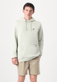 Helle grüne Kapuzenjacke aus weichem Stoff mit einer Fronttasche, Kordelzügen und einem kleinen Logo-Akzent auf der linken Brust. Getragen mit beigen Shorts.
