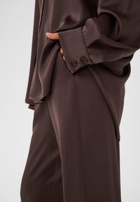 Ruskea silkkipaita pitkällä hihalla, jossa on rintatasku ja nappikiinnitys hihoissa, yhdistettynä matching ruskeisiin housuihin, sileä pinta.
