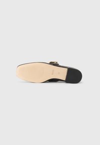 Black leather shoe featuring a square toe, gold-tone hardware, and a contrasting beige rubber sole with a textured grip pattern.
