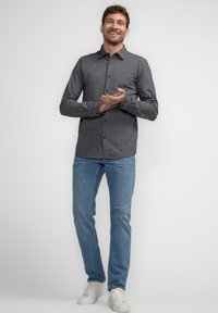 Skræddersyet skjorte i mørkegrå med hvide striber, parret med lyseblå jeans og hvide sneakers. Modellen står med åbne hænder.