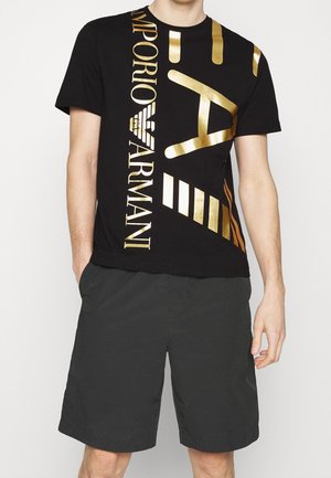 Mężczyzna w czarnych szortach i czarnej koszulce z dużym złotym logo i wzorem Emporio Armani na przodzie.