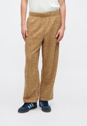 Person trägt braune, strukturierte, weit geschnittene Fleece-Hosen und dunkelblaue Sneaker mit hellblauen Streifen, steht vor einem einfarbigen Hintergrund.