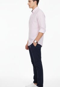 Hombre de perfil, vestido con una camisa rosa claro de estampado, mangas remangadas, pantalones azul marino oscuro, zapatillas blancas y un reloj de pulsera.