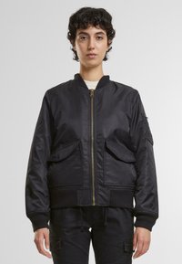Schwarze Bomberjacke mit frontaler Reißverschluss, gerippten Bündchen und zwei großen Fronttaschen. Glattes Material mit leichtem Glanz und klassischer Passform.