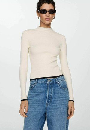 Top long-sleeve crème à côtes avec col montant, finition noire, associé à un jean en denim bleu taille haute, avec un délavage léger et une coupe relaxée.