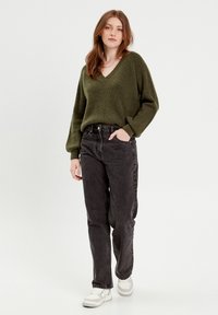 Pull en maille épaisse couleur olive avec un col en V. Associé à un jean noir taille haute. Modèle portant des baskets blanches à semelles texturées.