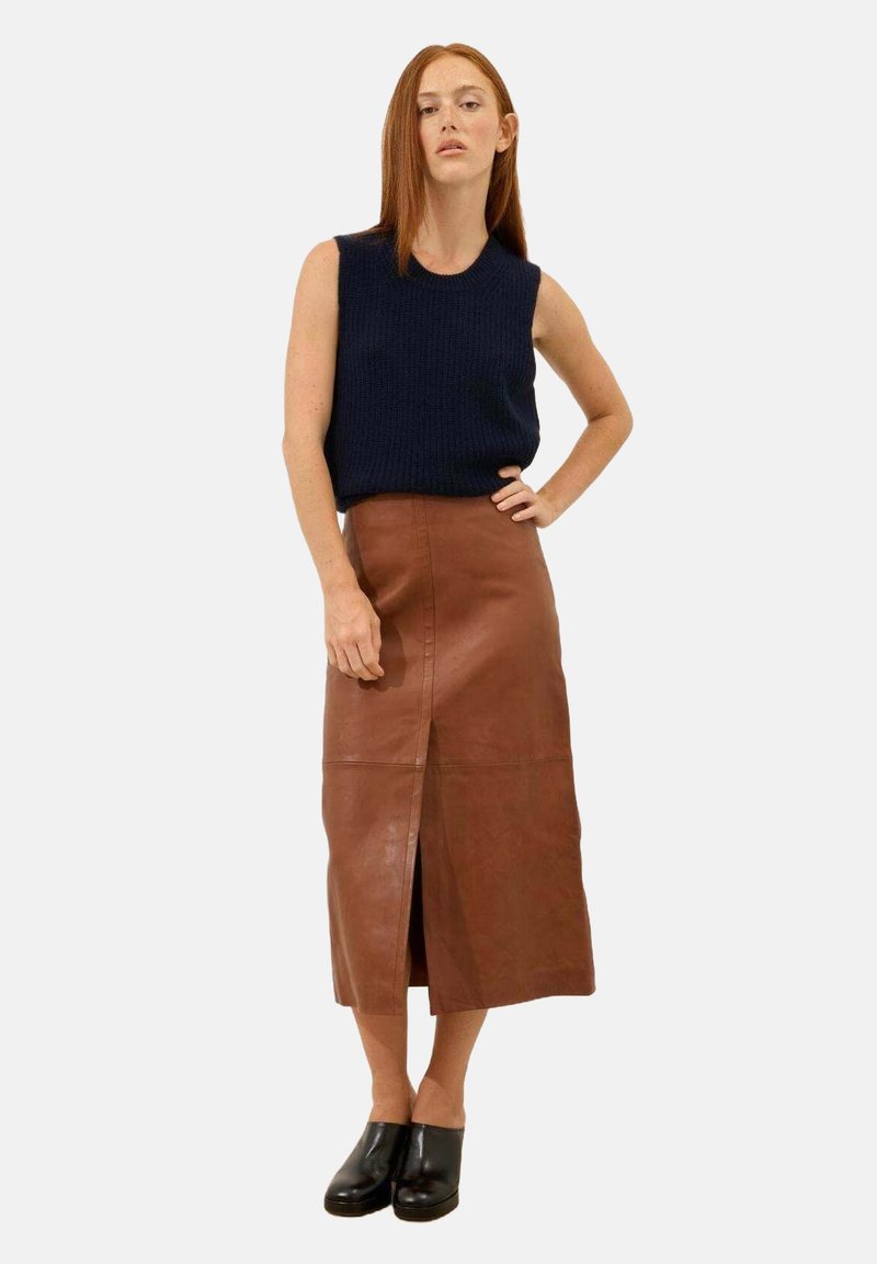 Falda midi de cuero marrón con una abertura frontal, combinada con una blusa de punto sin mangas de color azul oscuro; la modelo se encuentra de pie con una mano en la cadera.
