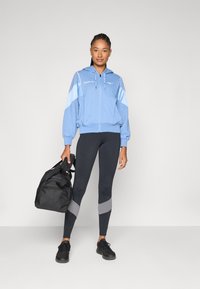 Um moletom azul claro com fecho, com detalhes cinzentos, combinado com leggings pretas com riscas cinzentas e sapatilhas pretas. Saco de desporto preto segurado na mão.