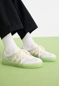Personne portant des baskets Adidas Samba blanches avec des semelles translucides vert pâle, des chaussettes blanches et un pantalon noir, debout sur un sol vert clair.