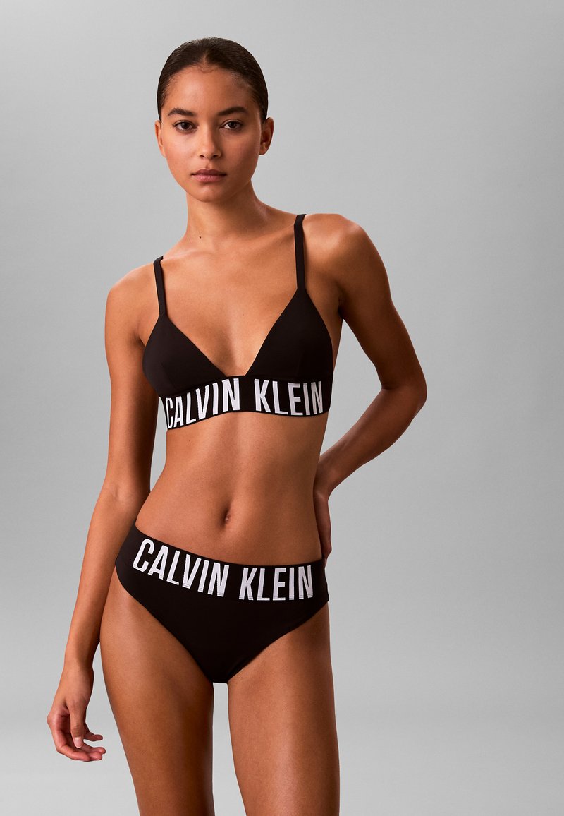 Zwart bikini set met een minimalistisch ontwerp en dunne bandjes. De band en tailleband tonen een prominent wit "CALVIN KLEIN" logo.