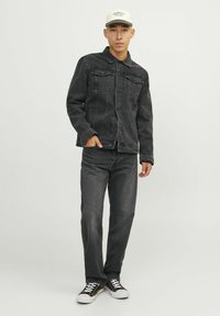 Svart denimjacka med två bröstfickor, mörka jeans och svarta sneakers. Modell med en ljus hatt, stående på en enhetlig bakgrund.