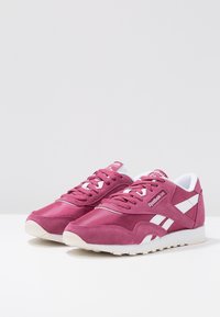 Růžové semišové a textilní tenisky s bílými akcenty a šněrováním, vybavené polstrovaným jazykem a texturovanou gumovou podešví. Logo Reebok na patě.