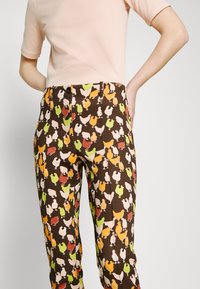 Pantalons à motifs colorés présentant un mélange de poules en orange, vert et crème sur un fond marron, avec une coupe classique et des poches avant.