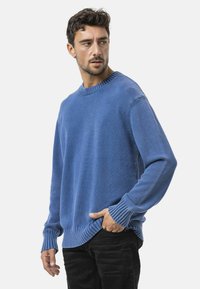 Blauwe gebreide trui met ribbelmanchetten en -zoom, met een klassiek ronde hals ontwerp. Soepele textuur, relaxte pasvorm, gedragen over zwarte broek.