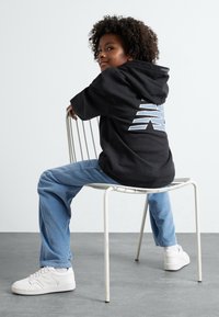 Felpa nera con cappuccio e un design geometrico blu sul retro, abbinata a pantaloni in velluto a coste blu chiaro e sneakers bianche.