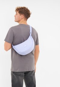 Világoskék, puha anyagból készült sling táska, ívelt formával, állítható pánttal és egy kis márkacímkével elöl.