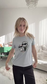 Blonde woman smiling, wearing light gray T-shirt with cartoon of a man and cat in sunglasses, standing in bright living room with white sofa.