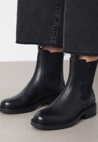 Botas de tobillo de cuero negro con punta redonda y paneles laterales elásticos. Suelas de goma negra gruesas. Llevadas con unos vaqueros oscuros cortos.