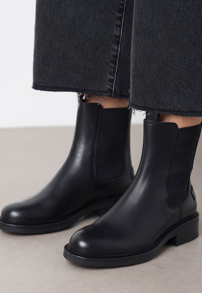 Botas de tobillo de cuero negro con punta redonda y paneles laterales elásticos. Suelas de goma negra gruesas. Llevadas con unos vaqueros oscuros cortos.