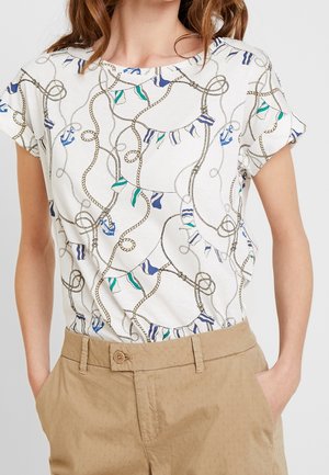 Kvinde iført en hvid kortærmet top med marineblå reb- og flagmønstre, stoppet ned i lysebrune bukser med høj talje og forlommer.