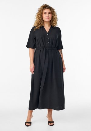 Kvinde med krøllet hår iført en sort midi-kjole med knapper, korte ærmer, bælte i taljen og sorte åbne sko med hæl, stående foran en hvid baggrund.